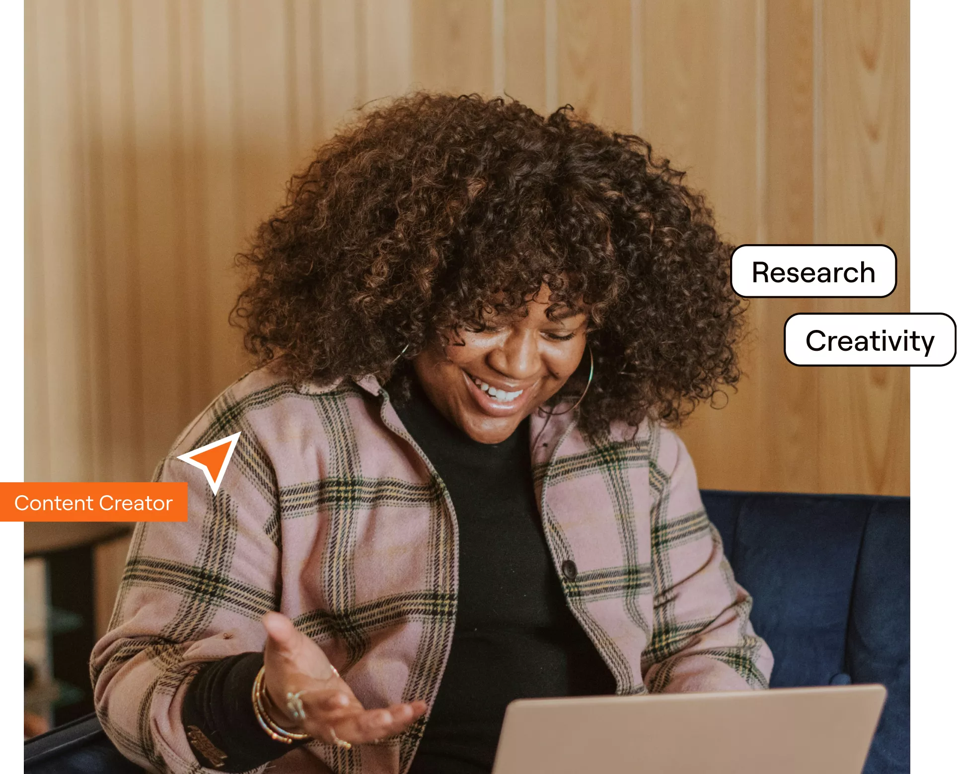 A woman with curly hair wearing a plaid shirt is seated on a couch, engaged in work on her laptop.
