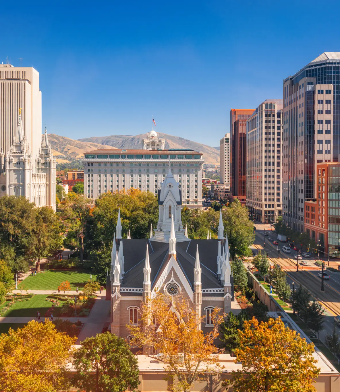 Cityscape view of Salt Lake City featuring historic LDS Church buildings, skyscrapers, and tree-lined streets with mountains in the background under a clear blue sky.