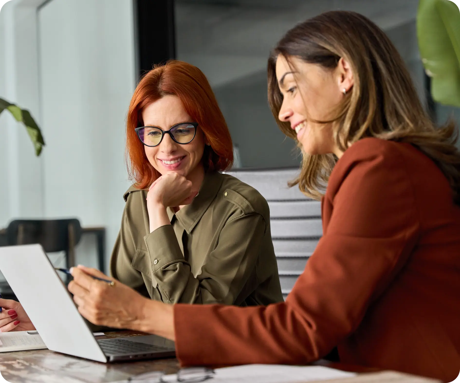 Two women in business attire collaborate at a table, focused on a laptop during a professional meeting.